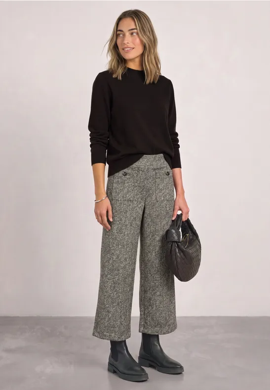 Frau in schwarzem Pullover, grauer Culotte, Boots und mit schwarzer Handtasche vor schlichtem Hintergrund.