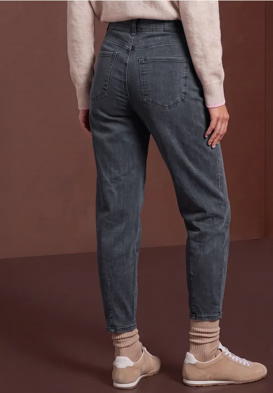 Rückansicht einer Person in hochgeschnittener grauer Jeans mit beigen Socken und Sneakers vor braunem Hintergrund.