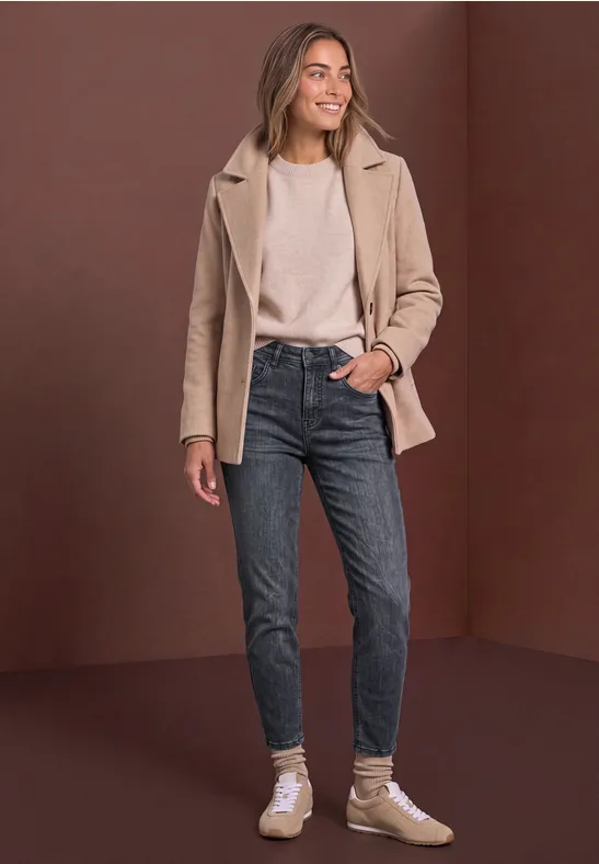 Frau in beigem Mantel, weißem Shirt, dunklen Jeans und Sneakers vor braunem Hintergrund.