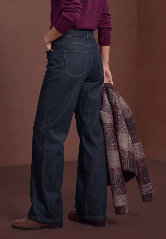 Eine von hinten fotografierte Frau trägt dunkle, weite Jeans, braune Schuhe und hält eine karierte Jacke.