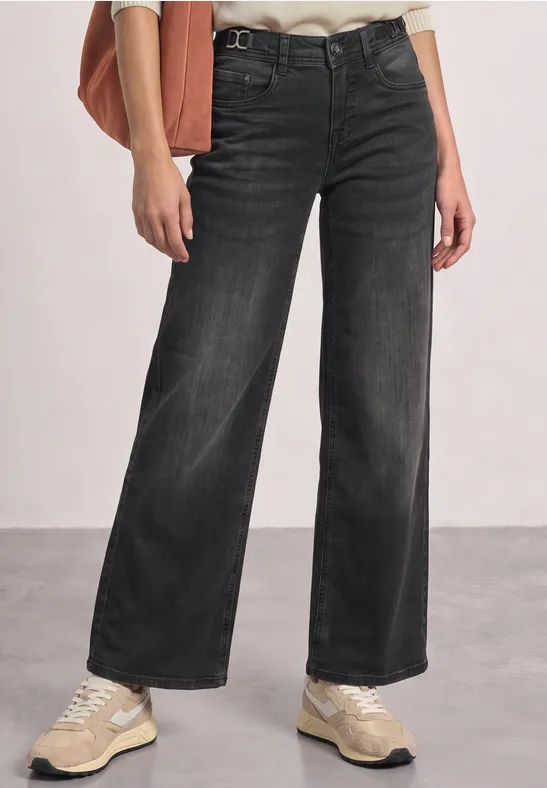 Schwarz-graue, weit geschnittene Jeans mit hellen Sneakern vor neutralem Hintergrund.