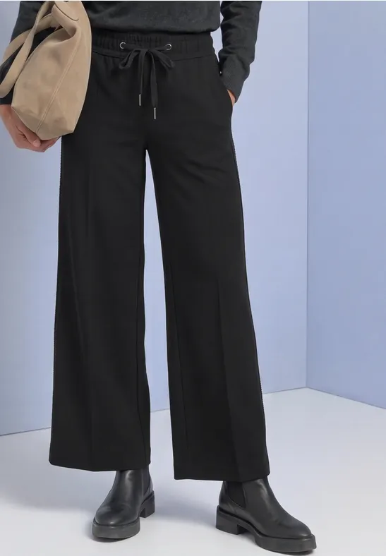 Zwarte, wijdvallende broek met aantrekkoord, zwarte laarzen en beige tas voor een lichtblauwe achtergrond.