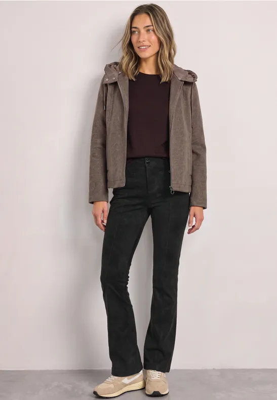 Frau trägt braunen Hoodie, dunkelrotes Shirt, schwarze Jeans und helle Sneaker vor hellem Hintergrund.