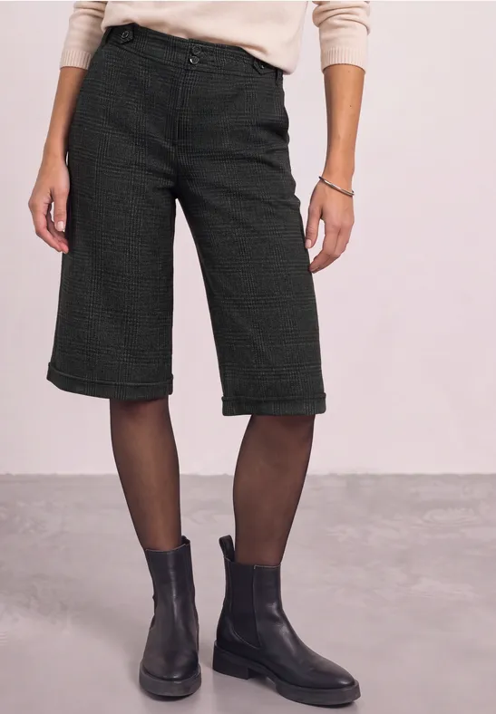 Person trägt schwarze Bermudashorts, feine Strumpfhosen und schwarze Stiefeletten vor hellem Hintergrund.