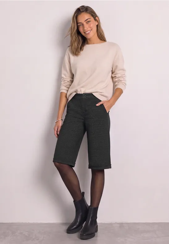 Frau trägt beigefarbenen Pullover, schwarze Bermuda-Shorts, feine Strumpfhose und Stiefeletten.