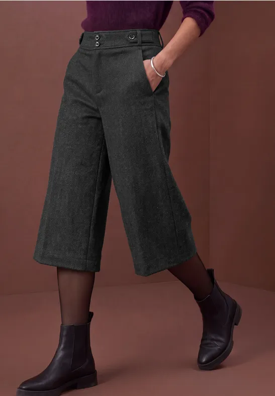 Pantalon culotte gris foncé, bottines noires et collants transparents sur fond rouge brique.