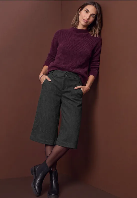 Femme en pull bordeaux et pantalon large noir devant un mur marron.
