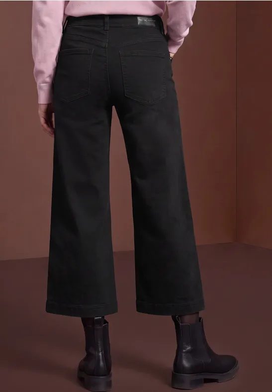 Vue de dos d’une personne en jean large noir et bottines noires sur fond marron.