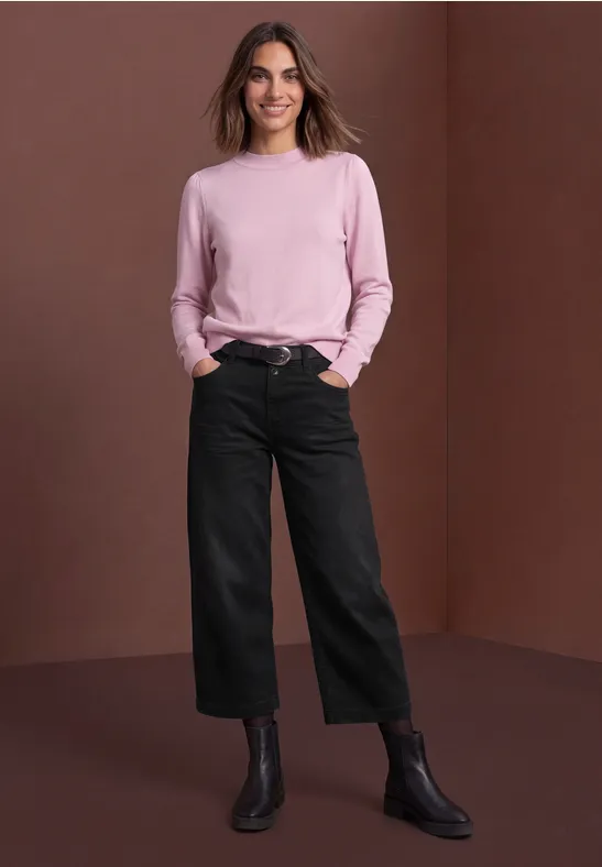 Femme souriante en pull rose, pantalon large noir et bottines, posant devant un fond marron.