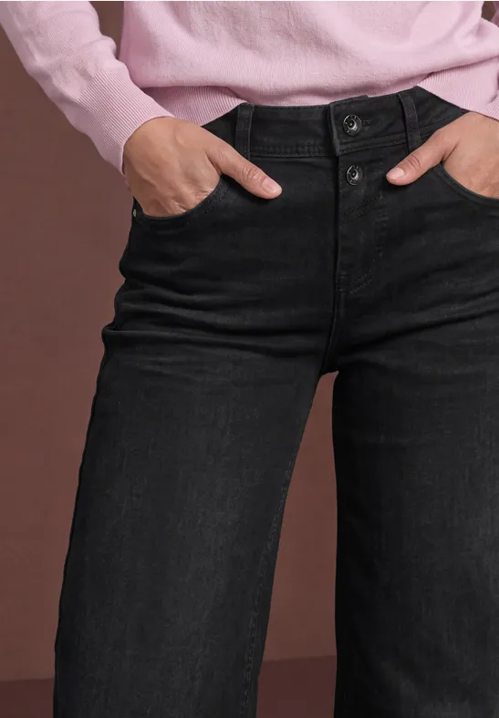Jean taille haute noir ample, mains dans les poches d'un pull rose.