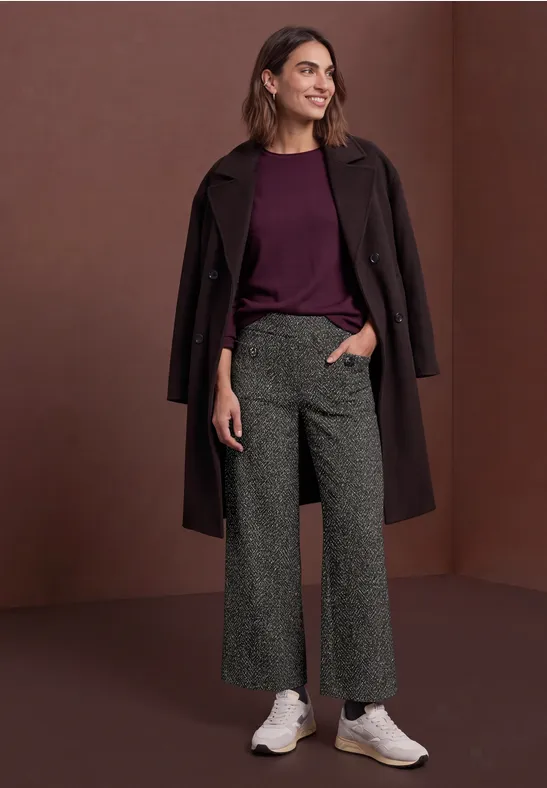 Frau trägt weinroten Pullover, dunklen Mantel, graue Culottes und weiße Sneaker vor schlichter brauner Wand.