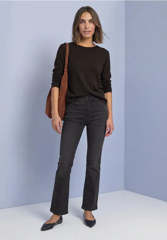 Frau in schwarzem Langarmshirt, schwarzen Bootcut-Jeans, Pumps und brauner Tasche vor pastellfarbenem Hintergrund.