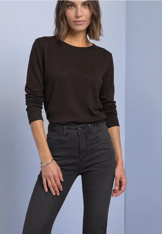 Frau in braunem Langarmshirt und dunkler Jeans steht vor neutralem Hintergrund.