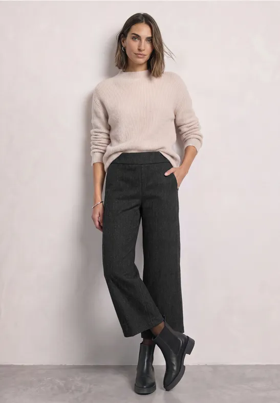Femme en pull clair, pantalon gris foncé ample et court, bottines noires, posant devant un mur clair.