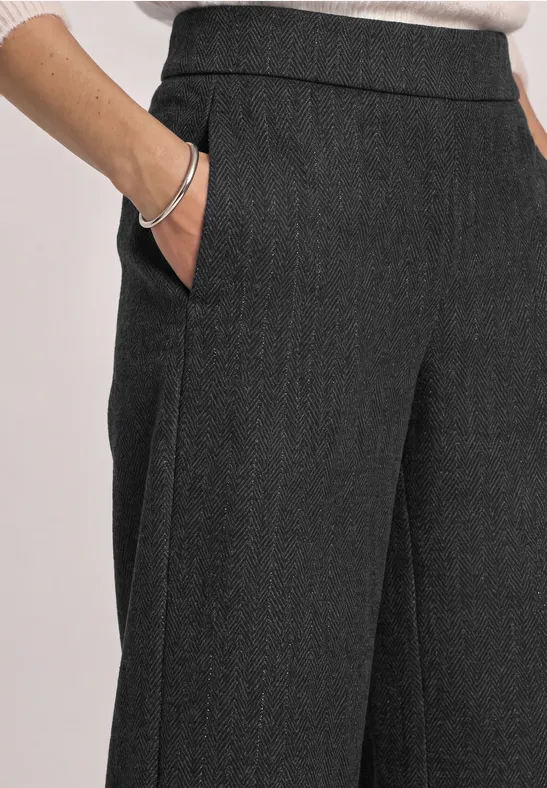 Pantalon en tissu gris foncé ample avec poches, main dans la poche, bracelet argenté visible.