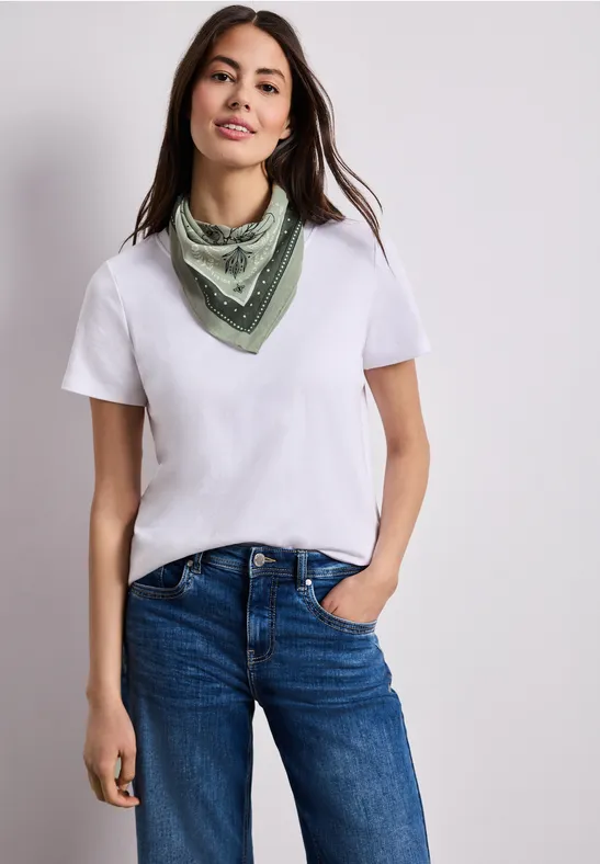 Frau mit weißem T-Shirt, blauer Jeans und grünem Halstuch, eine Hand in der Tasche.