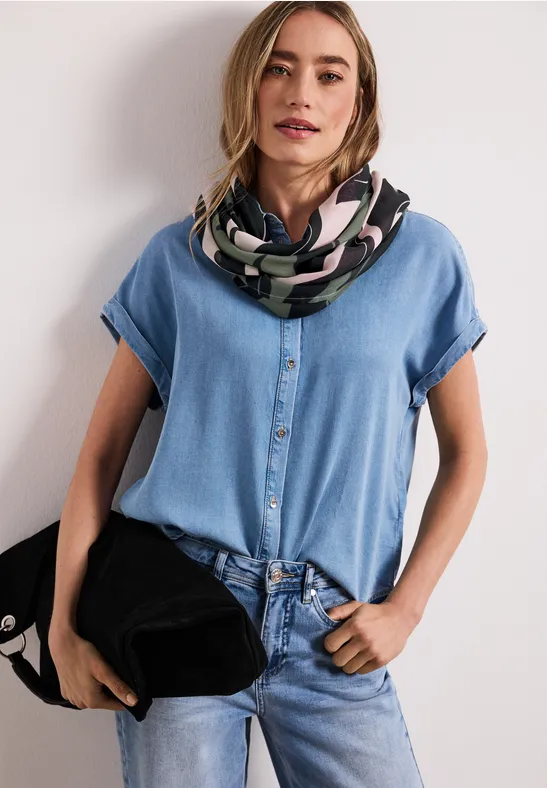 Kobieta w dżinsowej koszuli i spodniach, ma wzorzysty szalik i trzyma czarną torbę.