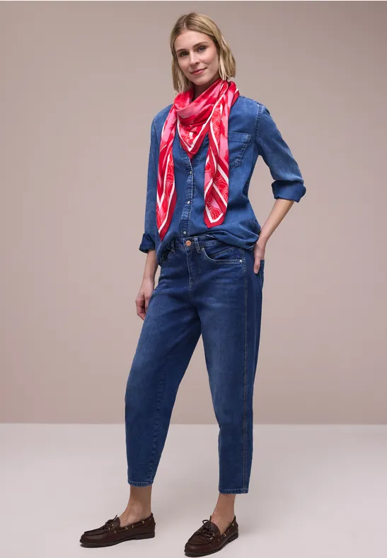 Kobieta nosi jeansową koszulę i spodnie, czerwona apaszka, brązowe mokasyny, casual look na neutralnym tle.