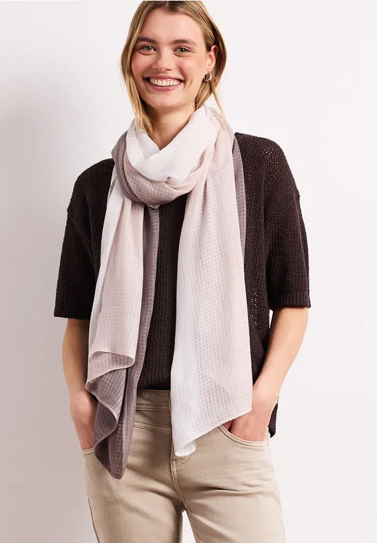 Kobieta w jasnym szaliku, czarnym kardiganie i beżowych spodniach, ręce w kieszeniach.