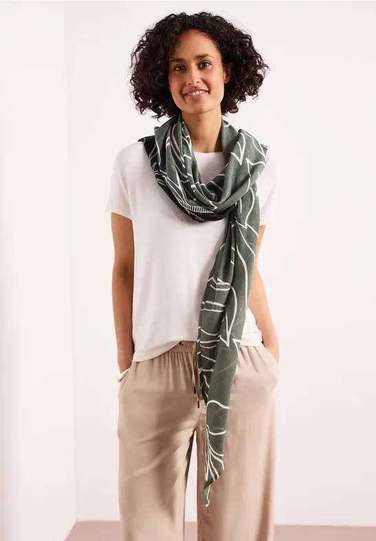 Osoba ma na sobie biały t-shirt, beżowe spodnie i zielony szalik na szyi.