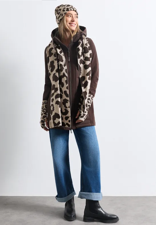 Eine Frau im Leoparden-Mützen-Set mit Schal und Handschuhen, braunem Mantel, weiter Jeans und schwarzen Boots.