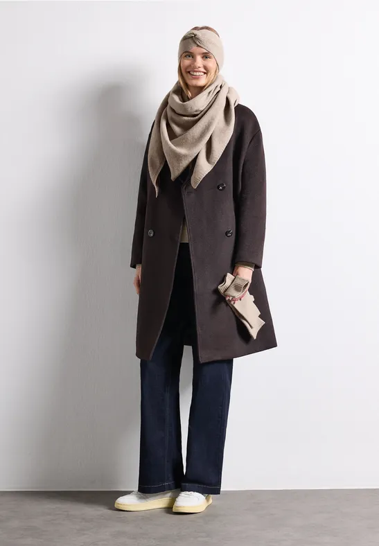 Eine Frau in dunklem Mantel, beigem Schal und weiten Jeans, hält eine Clutch, steht vor hellem Hintergrund.