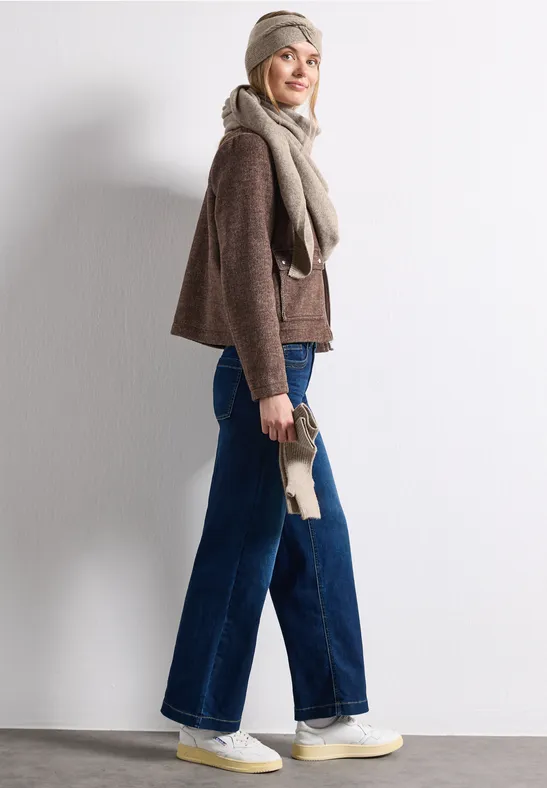 Frau in breiter Jeans, braunem Pullover, Schal und Mütze posiert seitlich vor weißem Hintergrund.