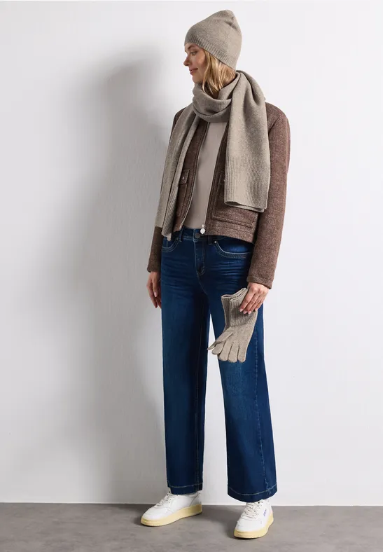 Frau in beige Mütze, Schal, braunem Pullover, Jeans, Handschuhen und weißen Sneakers vor weißem Hintergrund.