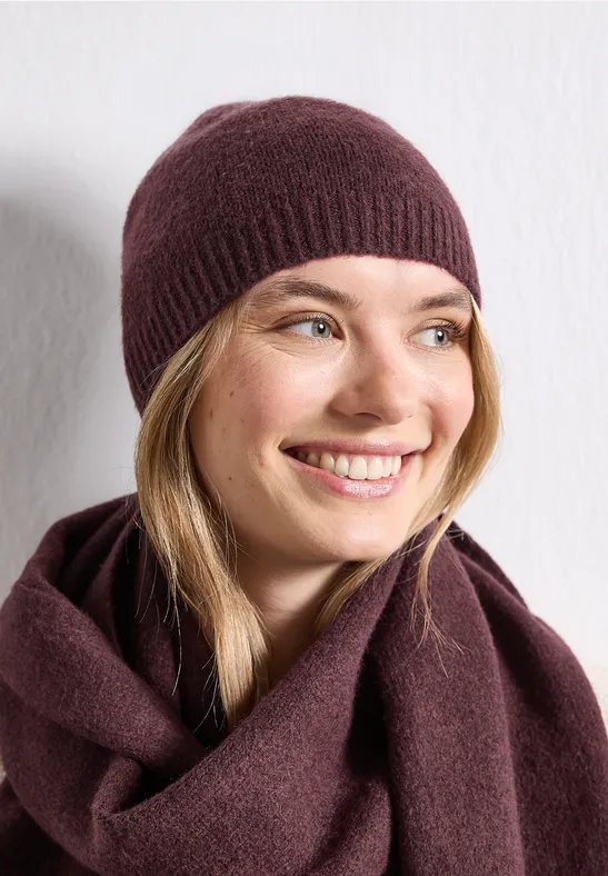Femme souriante portant un bonnet en maille bordeaux et une écharpe assortie, fond blanc.