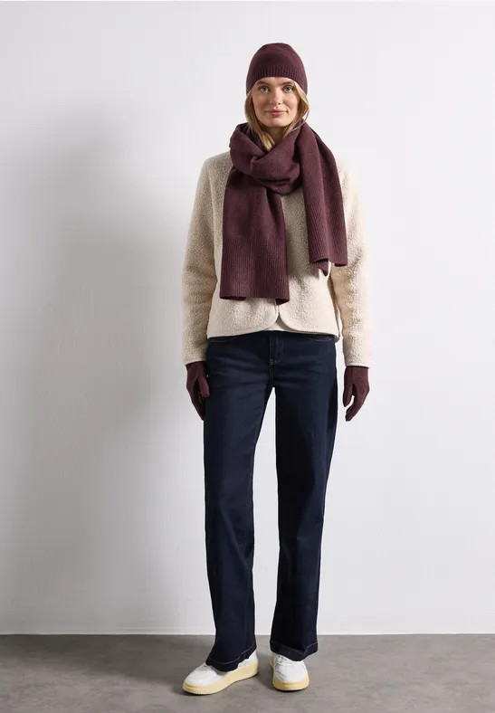 Personne debout devant un mur blanc, portant bonnet, écharpe, gants, pull clair et pantalon foncé.