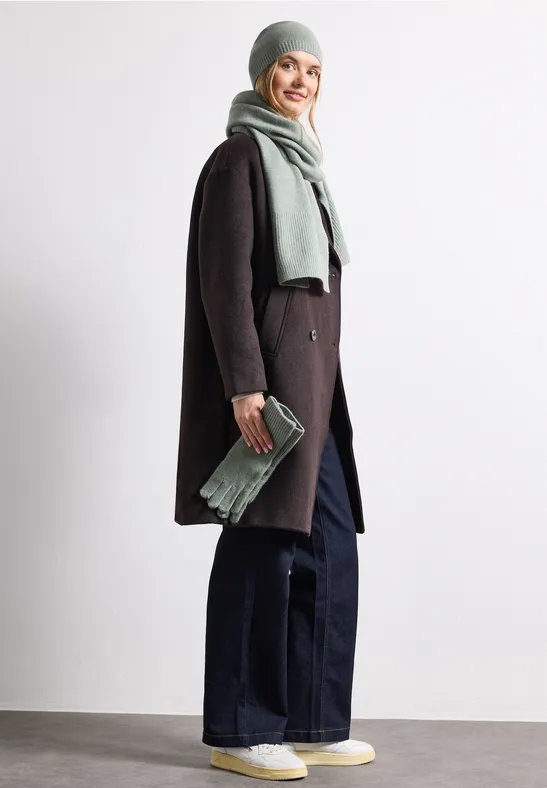 Frau mit Wintermantel, Schal und Mütze, weiten Jeans und Sneakern, lächelnd und hält Clutch