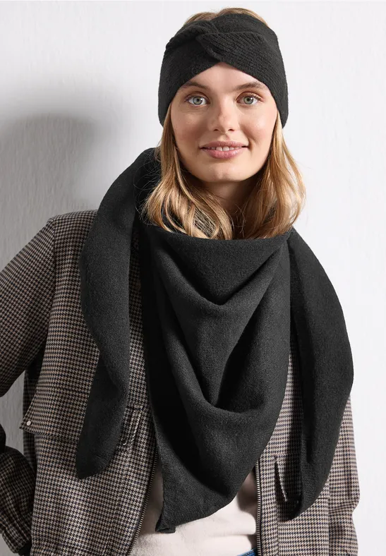 Frau mit schwarzem Stirnband und Dreieckstuch in schickem Karomantel, lächelnd vor heller Wand.