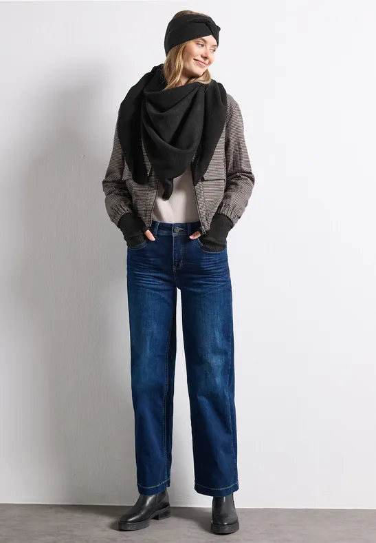 Frau in Jeans, grauer Jacke, schwarzem Schal und Mütze, steht vor weißem Hintergrund, lächelnd.