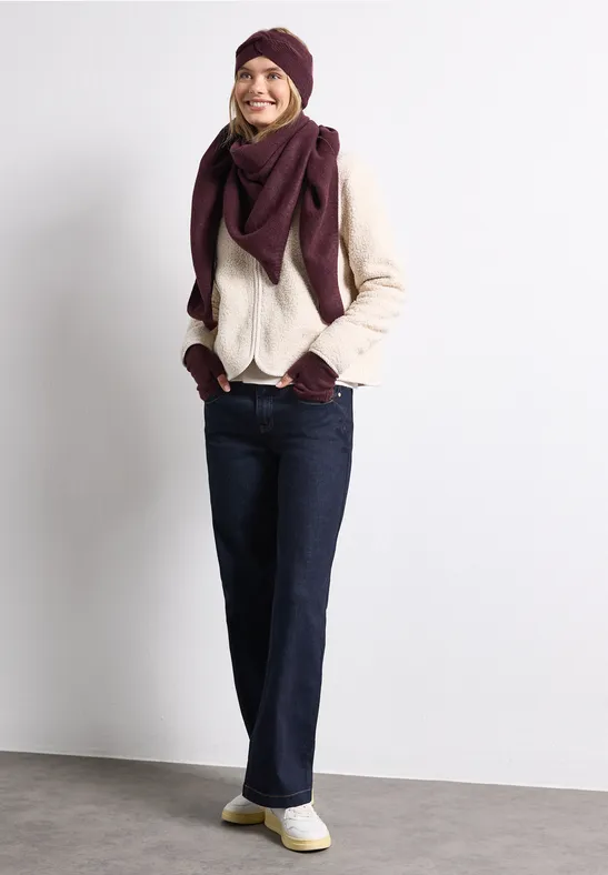 Femme souriante en tenue automnale : pull clair, bonnet et écharpe violets, jean foncé et baskets blanches.