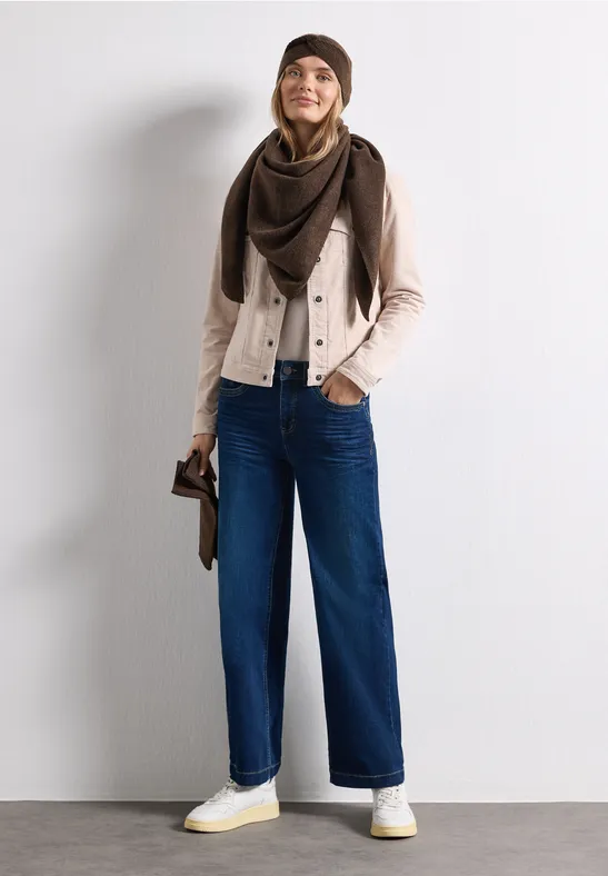 Eine junge Frau in Mütze, Schal, heller Jacke, weiten Jeans und weißen Sneakers lächelt.