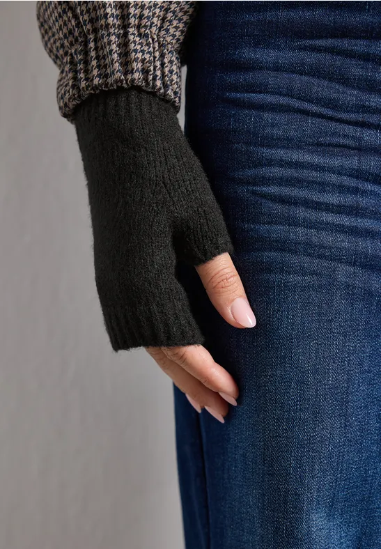 Schwarzer fingerloser Handschuh an einer Hand, kombiniert mit Jeans und Strickärmel im Hintergrund.