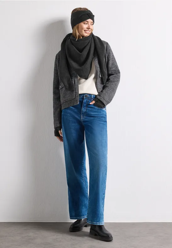Frau lehnt an weißer Wand, trägt Beanie, Schal, schwarze Jacke, weiße Bluse und weite blaue Jeans mit schwarzen Schuhen.