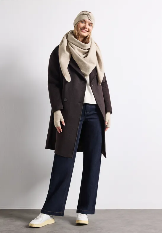 Frau in dunklem Mantel, weiter Jeans, cremefarbener Stola, Handschuhen und weißen Sneakers vor weißem Hintergrund.