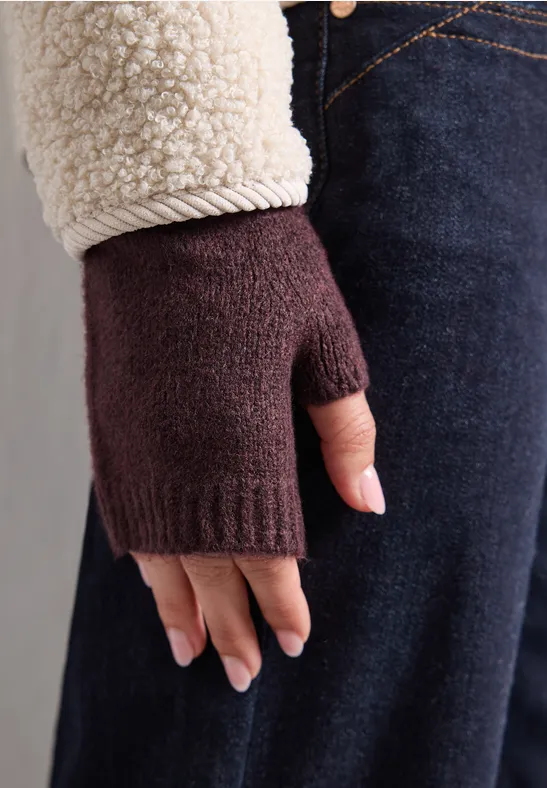 Hand mit burgunderrotem Strick-Fingerhandschuh, beigem Mantelärmel und dunklen Jeans