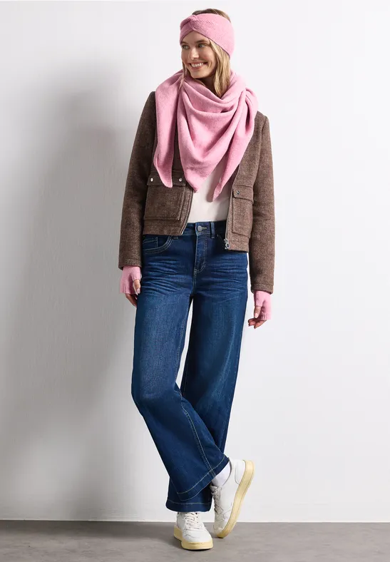 Frau mit pinkem Schal und Stirnband, brauner Jacke, weiten Jeans und weißen Sneakern vor weißem Hintergrund.