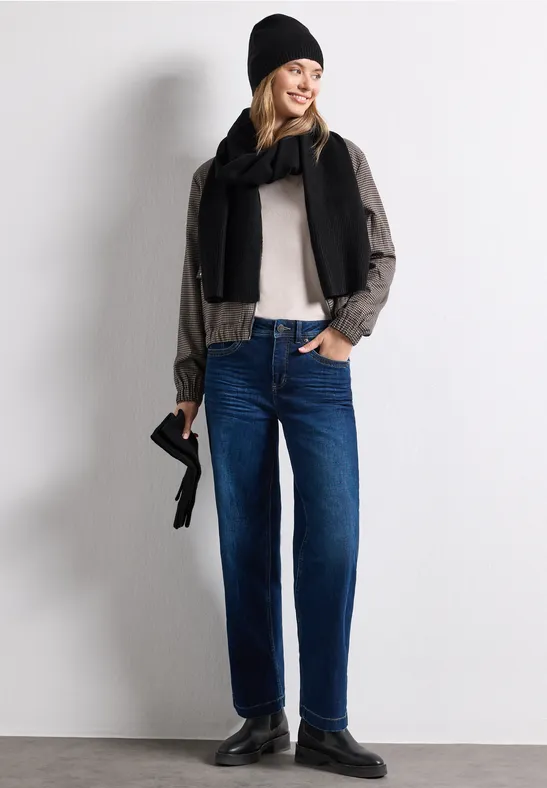 Frau in blauen Jeans, hellem Hemd, schwarzem Schal und Mütze sowie Loafers hält kleine Tasche vor grauem Hintergrund