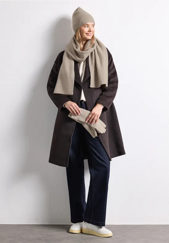 Femme en manteau long brun, bonnet et écharpe beiges, pantalon foncé et baskets blanches.