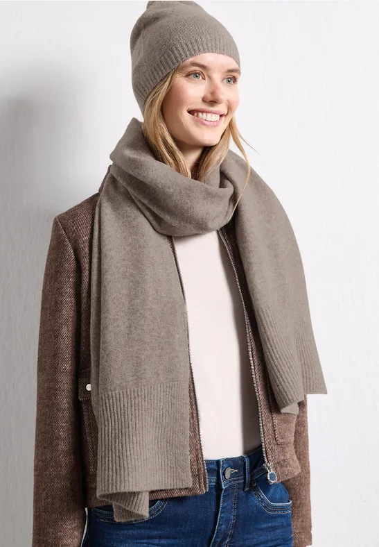Frau in beiger Mütze und Schal, brauner Strickjacke und Jeans, lächelnd vor weißer Wand.
