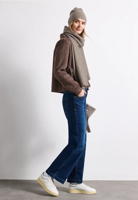 Eine Frau trägt braune Jacke, Schal, blaue Jeans und weiße Sneaker und steht seitlich lächelnd.