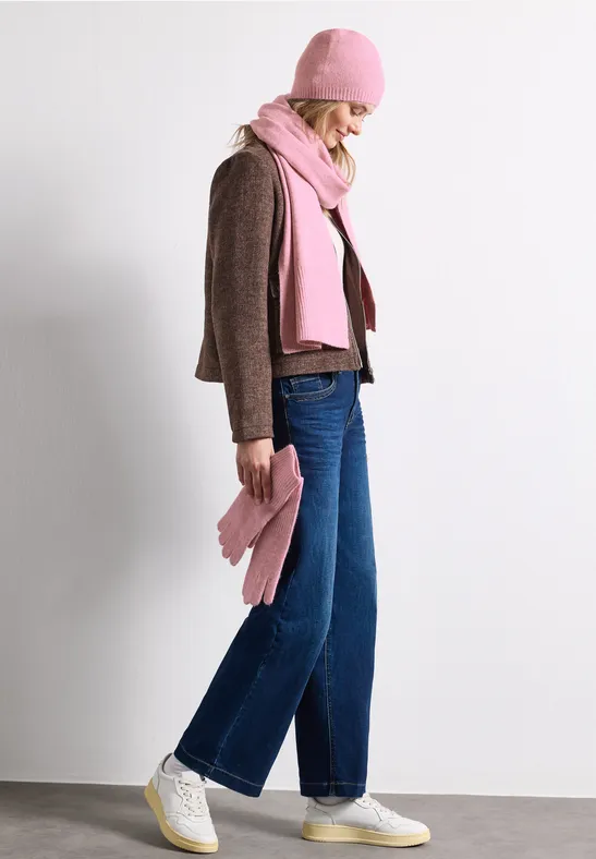 Frau im Profil, trägt braune Jacke, blaue Jeans, weiße Sneaker und pinke Mütze, Schal und Tasche.
