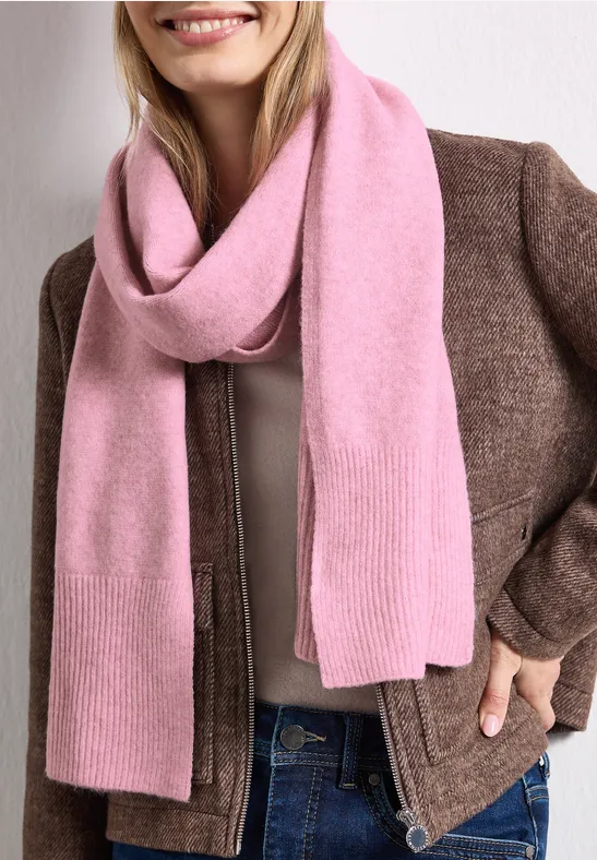 Eine lächelnde Frau mit rosafarbenem Schal, brauner Jacke und Jeans.
