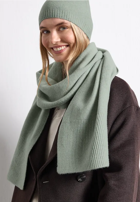 Eine lächelnde Frau trägt eine grüne Strickmütze und einen passenden Schal zu einem dunklen Mantel.