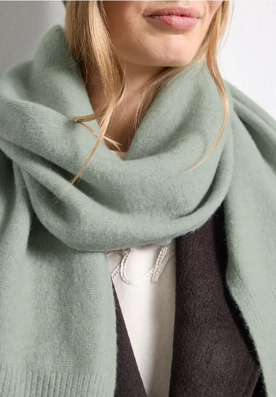 Eine Frau trägt einen weichen mintgrünen Schal und einen dunklen Mantel über einem hellen Pullover.