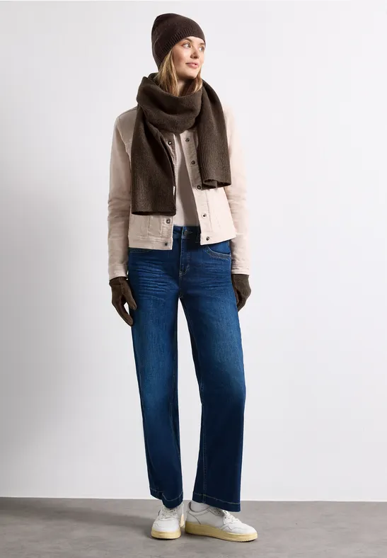 Frau trägt eine beige Jacke, blaue Jeans, braune Mütze, Schal, Handschuhe und weiße Sneaker.