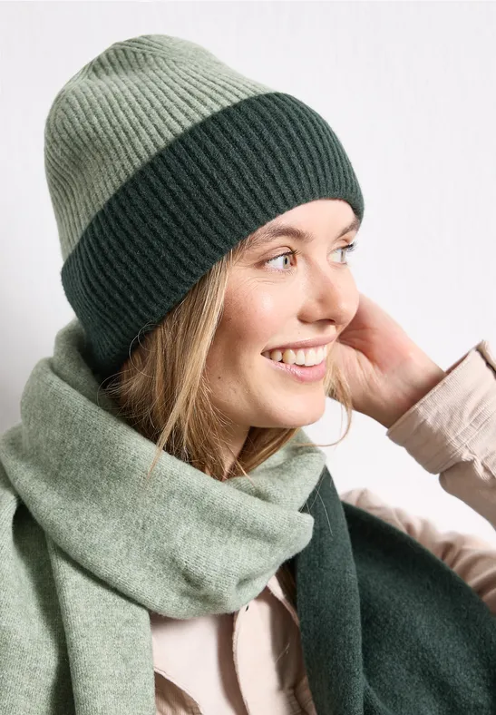 Frau mit zweifarbigem grünen Strickmütze und Schal, lächelnd.